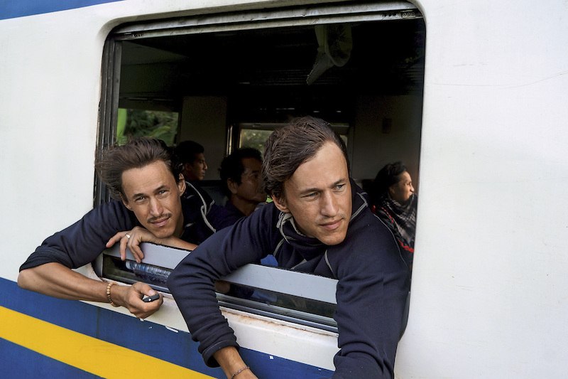 Two men lean out of a train window and look outside. Other passengers can be seen in the background., © Theaterhaus Stuttgart e.V. Two men lean out of a train window and look outside. Other passengers can be seen in the background., © Theaterhaus Stuttgart e.V.