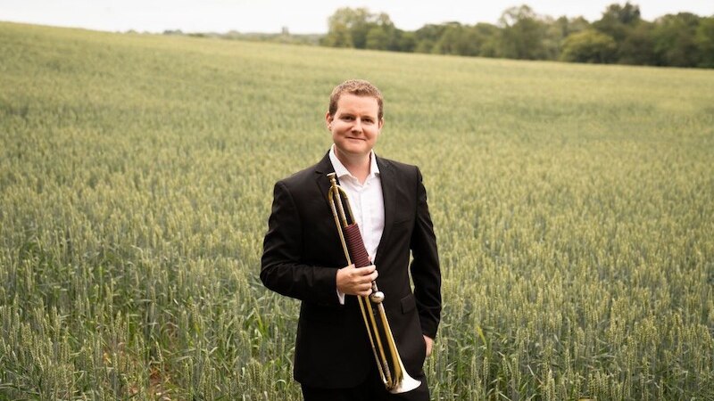 Ein Mann im Anzug steht in einem Weizenfeld und hält eine Trompete. Der Himmel ist bewölkt, und im Hintergrund sind Bäume zu sehen., © il Gusto Barocco - Stuttgarter Barockorchester