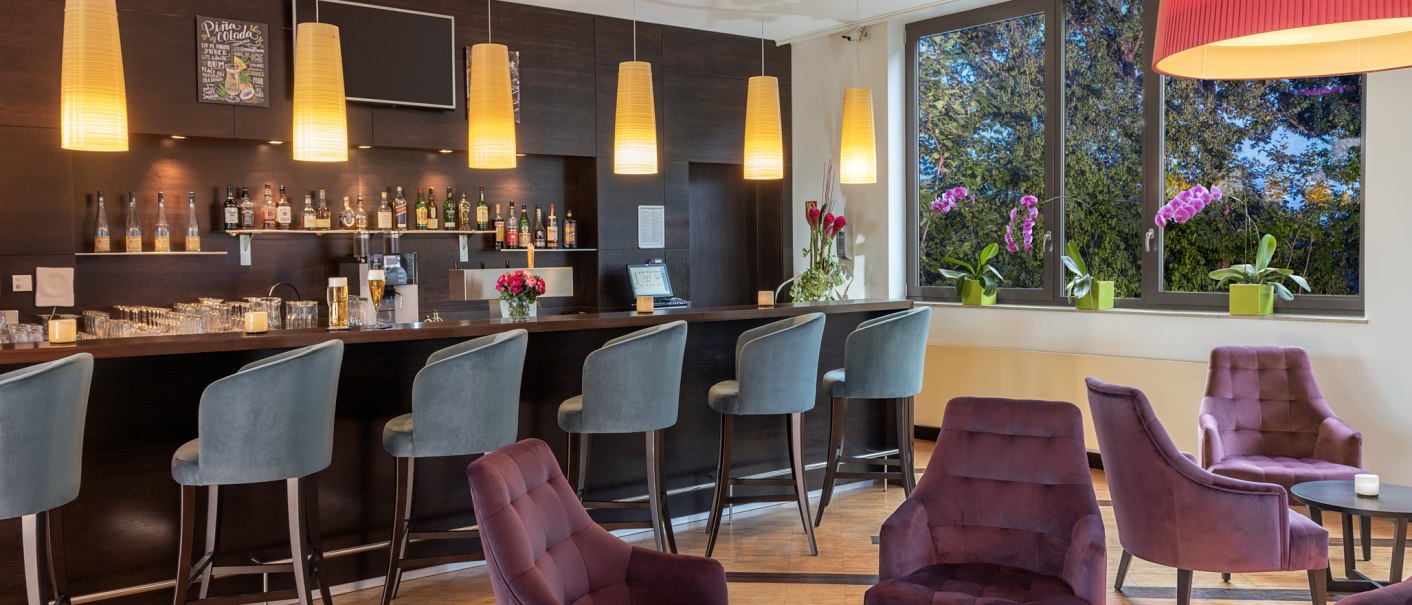 Elegante Bar mit lila Sesseln, Barhockern und h&auml;ngenden Lampen. Gro&szlig;e Fenster mit Blick auf B&auml;ume und Orchideen., &copy; NH Hotel Stuttgart Airport