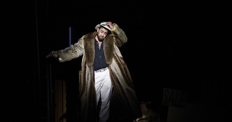 Ein Mann in einem langen Pelzmantel und Hut steht auf einer dunklen Bühne, hält eine Pistole und schaut in die Kamera., © Württembergische Staatstheater Stuttgart Ein Mann in einem langen Pelzmantel und Hut steht auf einer dunklen Bühne, hält eine Pistole und schaut in die Kamera., © Württembergische Staatstheater Stuttgart