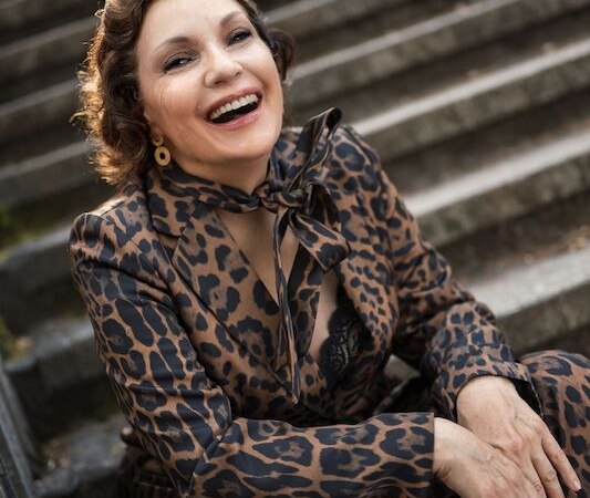 Eine Frau in einem Leopardenmuster-Anzug sitzt lächelnd auf einer Steintreppe und strahlt Freude aus., © Theaterhaus Stuttgart e.V. Eine Frau in einem Leopardenmuster-Anzug sitzt lächelnd auf einer Steintreppe und strahlt Freude aus., © Theaterhaus Stuttgart e.V.