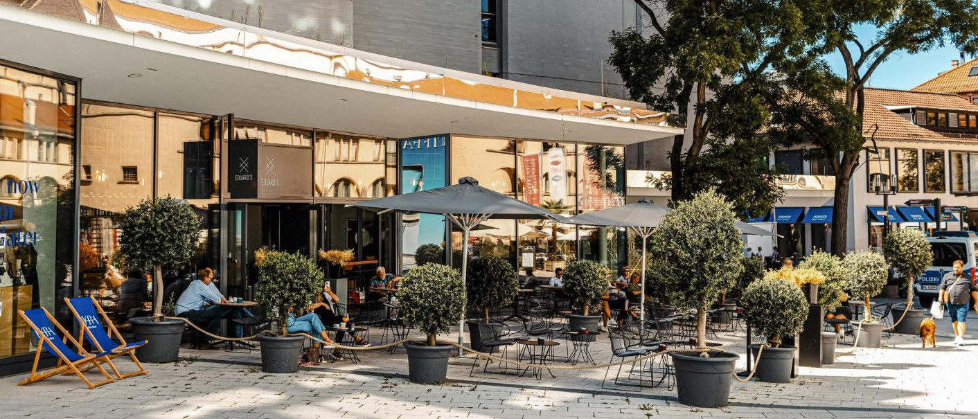 Außenbereich eines Cafés namens Eduard's mit Tischen, Stühlen und Sonnenschirmen. Menschen sitzen und entspannen, umgeben von Topfpflanzen., © SMG, Sarah Schmid