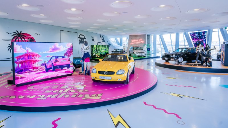 Colorful exhibition of classic cars, including a yellow Mercedes SLK. Neon lights and screens with retro designs decorate the room., © © Mercedes-Benz AG | Photo: Thomas Niedermüller