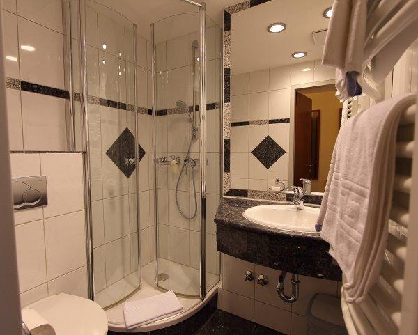 Modernes Badezimmer mit Glasdusche, Waschbecken und Handtuchhalter. Wei&szlig;e Fliesen mit schwarzen Akzenten. Beleuchtung an der Decke., &copy; Aparthotel Astro