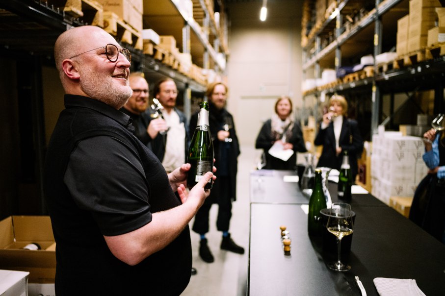 A group of people at a wine tasting in a storage room. One man is holding a wine bottle, while other participants are holding glasses., © Bernhard Kahrmann