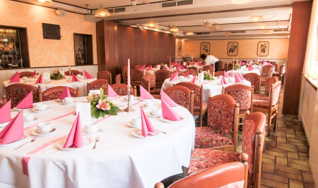Festlich gedeckte Tische in einem Restaurant mit rosa Servietten, Blumenarrangements und gedecktem Geschirr für eine Familienfeier., © Ulli Kellenbenz