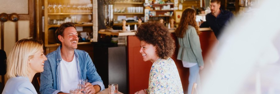 Three people sit laughing at a table in a cozy bar. Glasses and bottles can be seen in the background., © © Stuttgart-Marketing GmbH, Alwin Maigler Three people sit laughing at a table in a cozy bar. Glasses and bottles can be seen in the background., © © Stuttgart-Marketing GmbH, Alwin Maigler