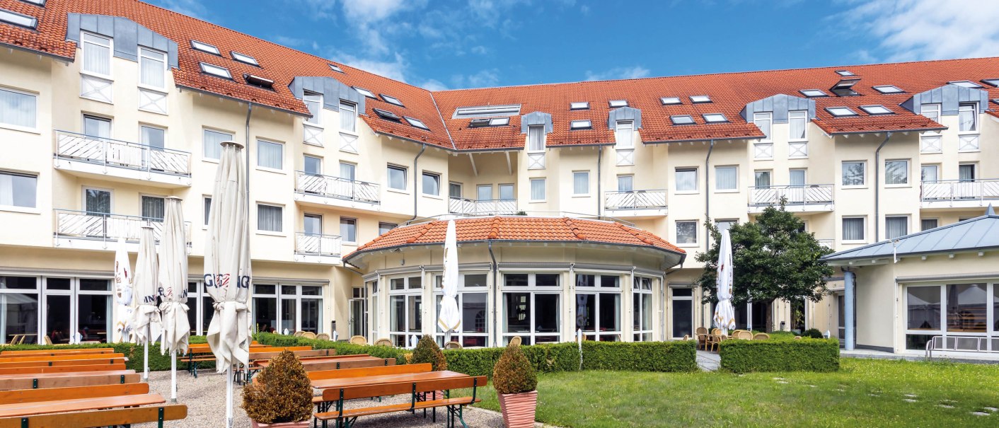 Hotel mit rotem Dach, Biergarten mit Holzbänken und geschlossenen Sonnenschirmen, umgeben von grüner Wiese und blühenden Sträuchern., © Seminaris Hotel Bad Boll