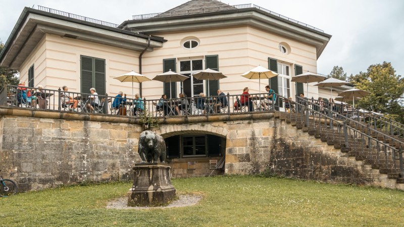 Das Bärenschlössle mit Terrasse, auf der Menschen unter Sonnenschirmen sitzen. Eine Bärenstatue steht im Vordergrund auf einer Wiese., © SMG, Sarah Schmid Das Bärenschlössle mit Terrasse, auf der Menschen unter Sonnenschirmen sitzen. Eine Bärenstatue steht im Vordergrund auf einer Wiese., © SMG, Sarah Schmid