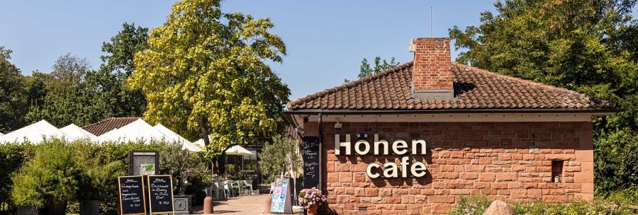 Das H&ouml;hencaf&eacute; im Killesbergpark zeigt ein Geb&auml;ude aus Backstein mit Terrasse, umgeben von B&auml;umen und Pflanzen. Wei&szlig;e Sonnenschirme sind sichtbar., &copy; Stuttgart-Marketing GmbH, Sarah Schmid