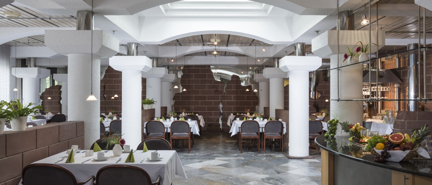 Stilvolles Frühstücksrestaurant mit weißen Tischdecken, Pflanzen und moderner Architektur. Ein Buffet mit frischem Obst ist im Vordergrund zu sehen., © Hotel Berlin aZIS Hotel BetriebsGmbH