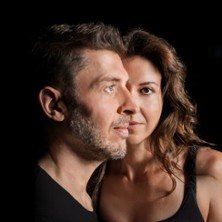 A man and a woman look into the distance against a black background. Both are wearing dark clothing., &copy; see on the left