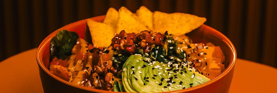 A bowl of avocado, sesame seeds, vegetables and potato chips stands on a table against a striped background., © Blackbook Studios A bowl of avocado, sesame seeds, vegetables and potato chips stands on a table against a striped background., © Blackbook Studios