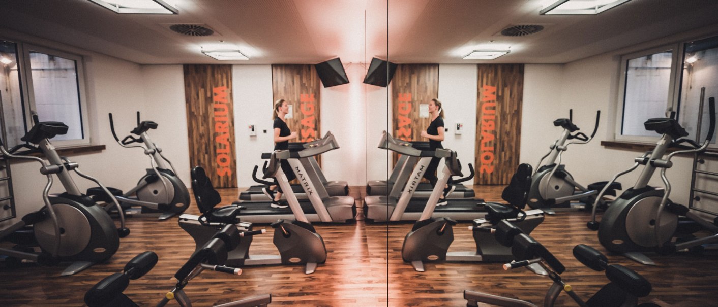 Ein Fitnessraum mit Spiegelwand, Laufband und verschiedenen Fitnessgeräten auf Holzboden. Eine Person trainiert auf dem Laufband., © Abacco Hotels GmbH Ein Fitnessraum mit Spiegelwand, Laufband und verschiedenen Fitnessgeräten auf Holzboden. Eine Person trainiert auf dem Laufband., © Abacco Hotels GmbH