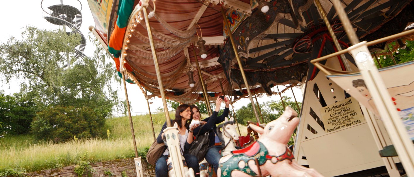 Zwei Personen fahren auf einem nostalgischen Karussell mit Pferdefiguren. Im Hintergrund ist ein spiralförmiger Aussichtsturm zu sehen., © SMG, Christoph Düpper