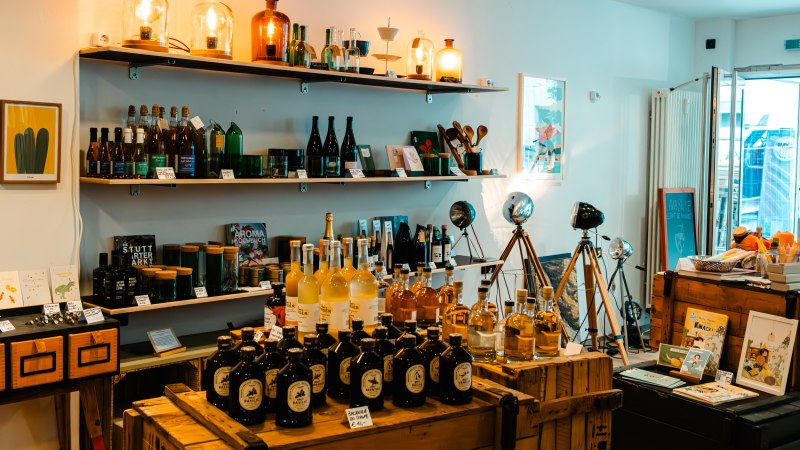 Ein gemütlicher Laden mit Regalen voller Flaschen, Lampen und Dekorationsartikeln. Holzkisten und Tripod-Lampen ergänzen das Ambiente., © SMG_Sarah Schmid Ein gemütlicher Laden mit Regalen voller Flaschen, Lampen und Dekorationsartikeln. Holzkisten und Tripod-Lampen ergänzen das Ambiente., © SMG_Sarah Schmid