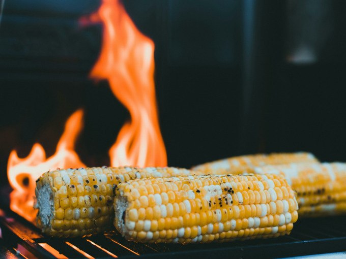 Drei Maiskolben liegen auf einem Grillrost, während im Hintergrund Flammen lodern. Perfekt für ein Veggie BBQ., © Cool-Tours StattReisen Drei Maiskolben liegen auf einem Grillrost, während im Hintergrund Flammen lodern. Perfekt für ein Veggie BBQ., © Cool-Tours StattReisen