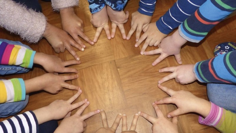 Kinderh&auml;nde in bunten &Auml;rmeln formen einen Kreis auf einem Holzboden. Die Finger zeigen zur Mitte und symbolisieren Gemeinschaft., &copy; Christian Steeneck