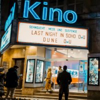 Entrance to a movie theater at night with illuminated sign. People stand in front of it and look up. Movies: 'Last Night in Soho', 'Dune'., © SMG, Alwin Maigler Entrance to a movie theater at night with illuminated sign. People stand in front of it and look up. Movies: 'Last Night in Soho', 'Dune'., © SMG, Alwin Maigler