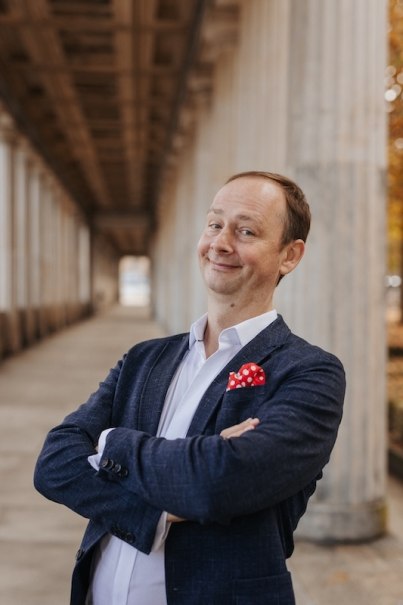 A man in a blue suit with a red handkerchief stands smiling in a portico., &copy; Renitenztheater Stuttgart e.V.