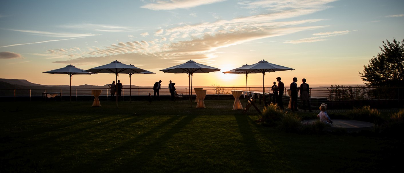 Hotelterrasse bei Sonnenuntergang, mehrere Sonnenschirme und Menschen im Schatten. Der Himmel ist mit Wolken bedeckt, die Sonne steht tief am Horizont., © Achalm. Hotel GmbH & Co. KG Hotelterrasse bei Sonnenuntergang, mehrere Sonnenschirme und Menschen im Schatten. Der Himmel ist mit Wolken bedeckt, die Sonne steht tief am Horizont., © Achalm. Hotel GmbH & Co. KG