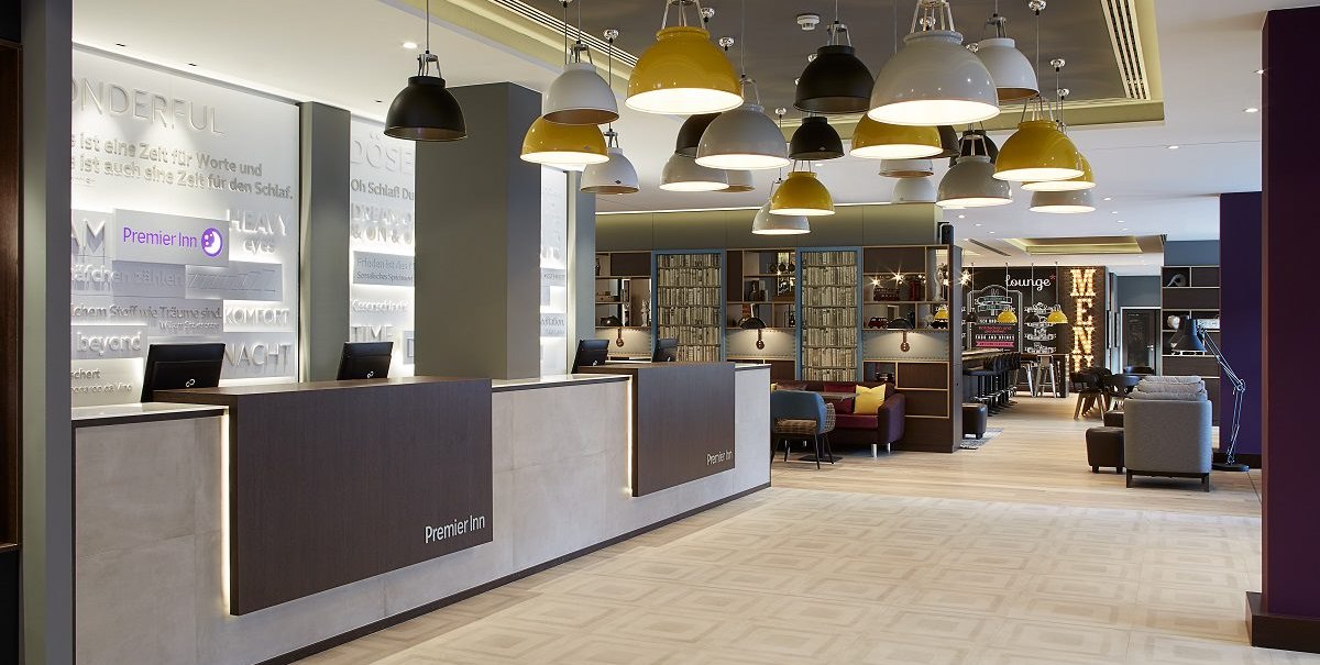Modern hotel lobby with reception, stylish hanging lamps and cozy seating area. An illuminated menu sign can be seen in the background., © Premier Inn Modern hotel lobby with reception, stylish hanging lamps and cozy seating area. An illuminated menu sign can be seen in the background., © Premier Inn