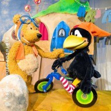 Der kleine Rabe Socke fährt auf einem Laufrad, während ein Bär ihm zuschaut. Im Hintergrund ist ein buntes Haus zu sehen., © links im Bild Der kleine Rabe Socke fährt auf einem Laufrad, während ein Bär ihm zuschaut. Im Hintergrund ist ein buntes Haus zu sehen., © links im Bild