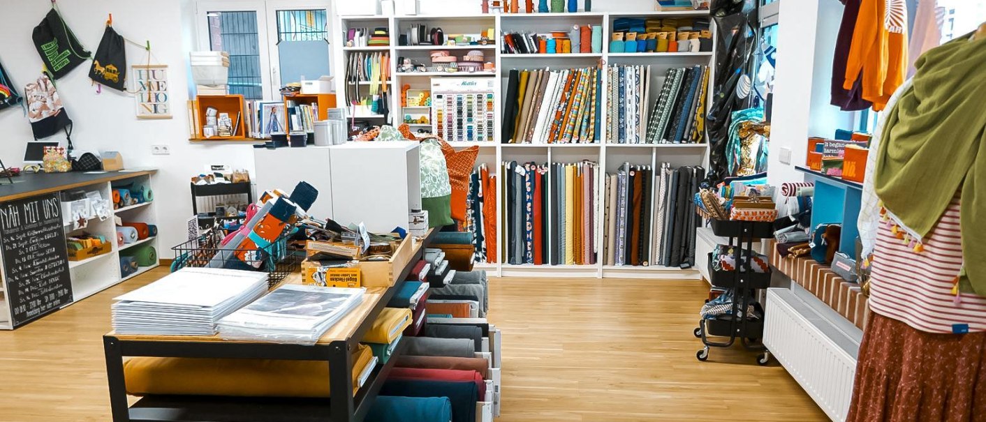Ein Stoffladen mit Regalen voller bunter Stoffrollen und Nähzubehör. Auf einem Tisch liegen weitere Materialien. An der Wand hängen Taschen., © Lena Beck, Hummelkonig GbR, Stuttgart