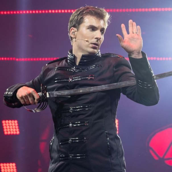 A man in a black jacket performs a sword illusion on a stage. He holds a sword and raises his hand as if he is performing magic., &copy; Renitenztheater Stuttgart e.V.