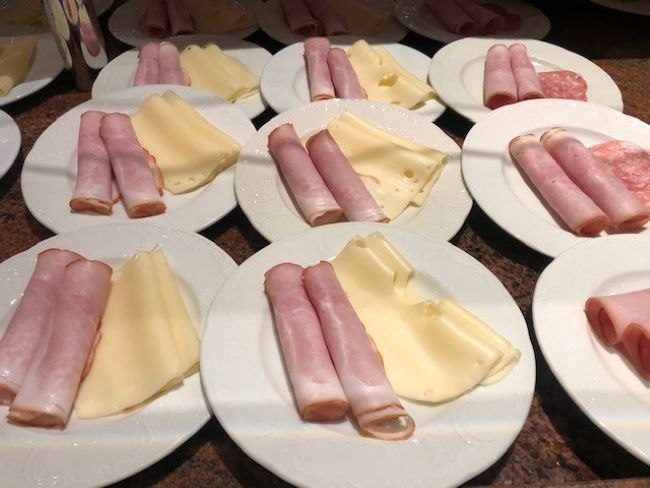 Weiße Teller mit aufgerolltem Schinken und Käsescheiben, auf einem Tisch arrangiert. Frühstücksvorbereitung., © Kronen Hotel GmbH Weiße Teller mit aufgerolltem Schinken und Käsescheiben, auf einem Tisch arrangiert. Frühstücksvorbereitung., © Kronen Hotel GmbH