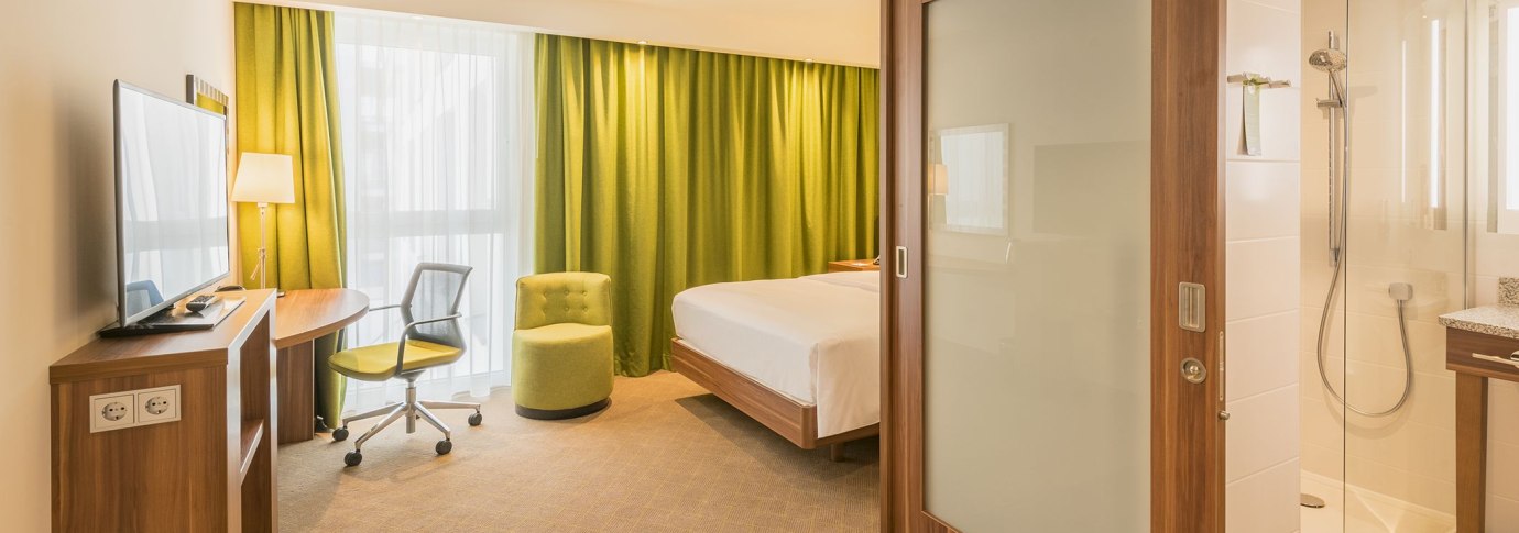 Modernes Hotelzimmer mit Doppelbett, gr&uuml;nem Sessel, Schreibtisch und Blick ins Badezimmer mit Dusche. Helle Vorh&auml;nge und gr&uuml;ne Gardinen., &copy; Hampton by Hilton Stuttgart City Centre