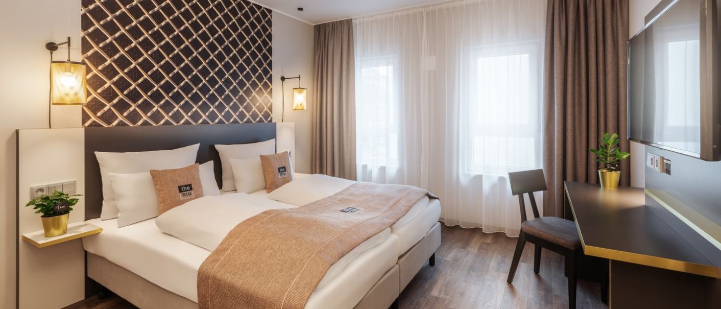A modern hotel room with a double bed, a decorative wall, a desk, and plants. Light-colored curtains and warm lighting create a cozy atmosphere., &copy; Novum Hospitality