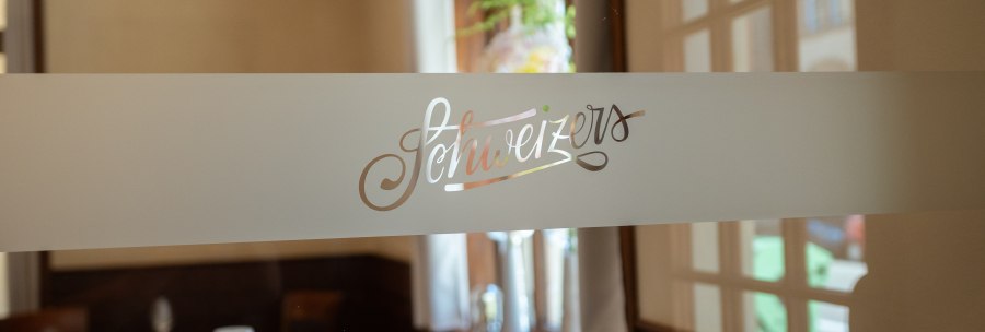 An elegant restaurant with the lettering 'Schweizers' on a glass door, tables set in the background., &copy; Schweizers Restaurant