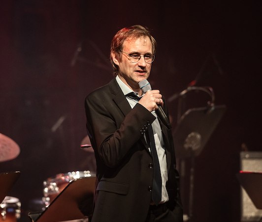 Ein Mann im Anzug spricht in ein Mikrofon auf einer B&uuml;hne. Im Hintergrund sind Musikinstrumente wie ein Schlagzeug und Notenst&auml;nder zu sehen., &copy; Staatliche Hochschule f&uuml;r Musik und Darstellende Kunst