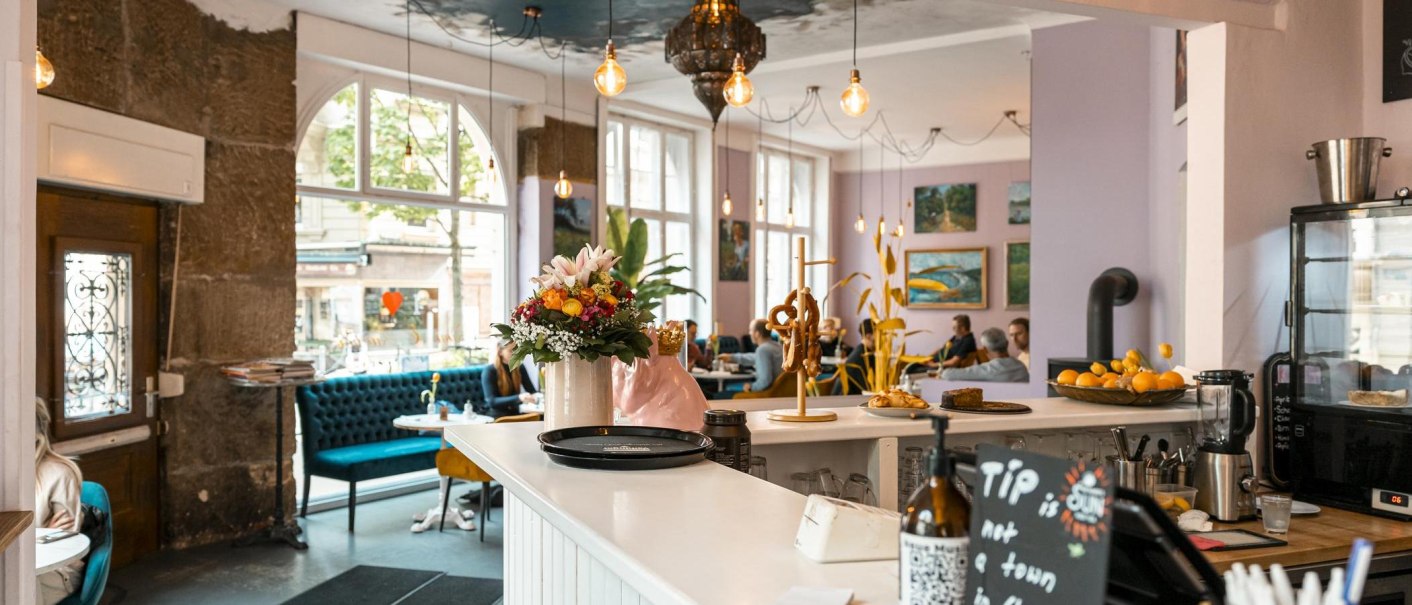 Cozy café with flowers and pretzels on the counter, blue sofas and art on the walls. Large windows let in plenty of light., © SMG Stuttgart Marketing GmbH - Sarah Schmid Cozy café with flowers and pretzels on the counter, blue sofas and art on the walls. Large windows let in plenty of light., © SMG Stuttgart Marketing GmbH - Sarah Schmid