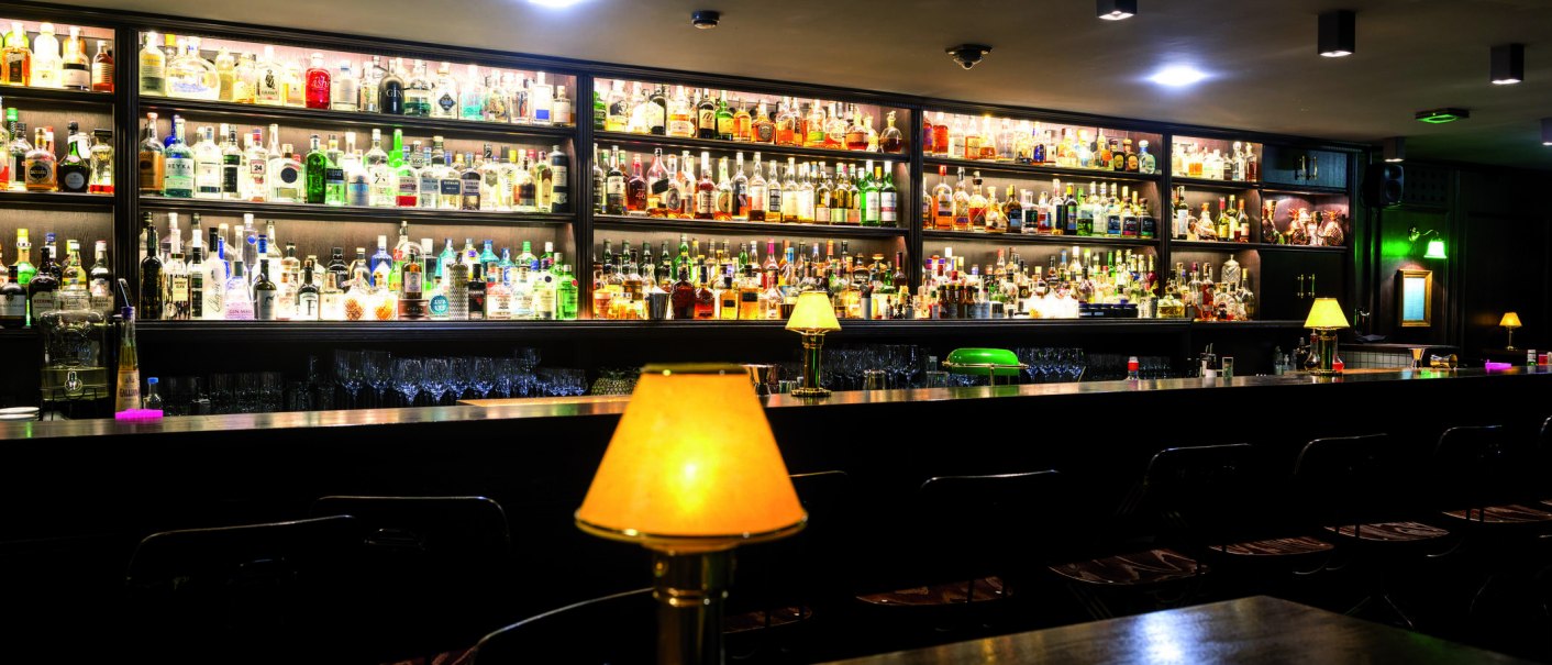 Eine stilvolle Bar mit zahlreichen Flaschen in Regalen und warm beleuchteten Lampen auf der Theke., © TMBW, Gregor Lengler Eine stilvolle Bar mit zahlreichen Flaschen in Regalen und warm beleuchteten Lampen auf der Theke., © TMBW, Gregor Lengler