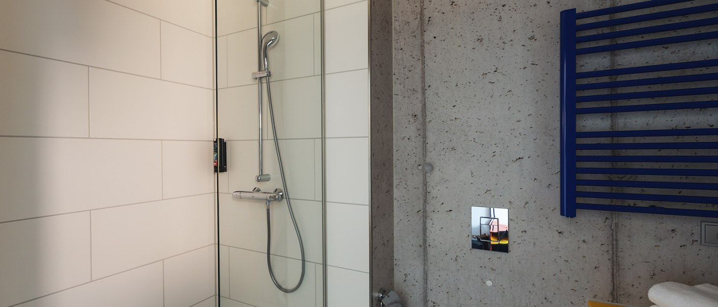 A modern bathroom with a glass shower, white tiles, a blue towel rack, and a yellow shelf with towels., © the niu Form - Novum Hospitality A modern bathroom with a glass shower, white tiles, a blue towel rack, and a yellow shelf with towels., © the niu Form - Novum Hospitality