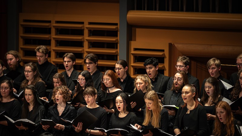 Ein Chor in schwarzer Kleidung singt auf einer B&uuml;hne. Die S&auml;ngerinnen und S&auml;nger halten Notenbl&auml;tter in den H&auml;nden., &copy; Staatliche Hochschule f&uuml;r Musik und Darstellende Kunst