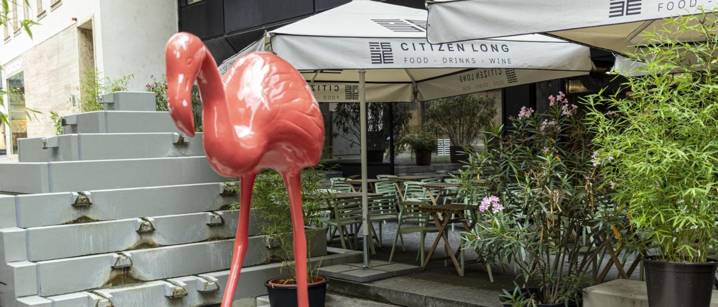 Ein großer, pinker Flamingo steht vor dem Café Citizen Long. Im Hintergrund sind Tische, Stühle und Pflanzen zu sehen., © SMG, Sarah Schmid