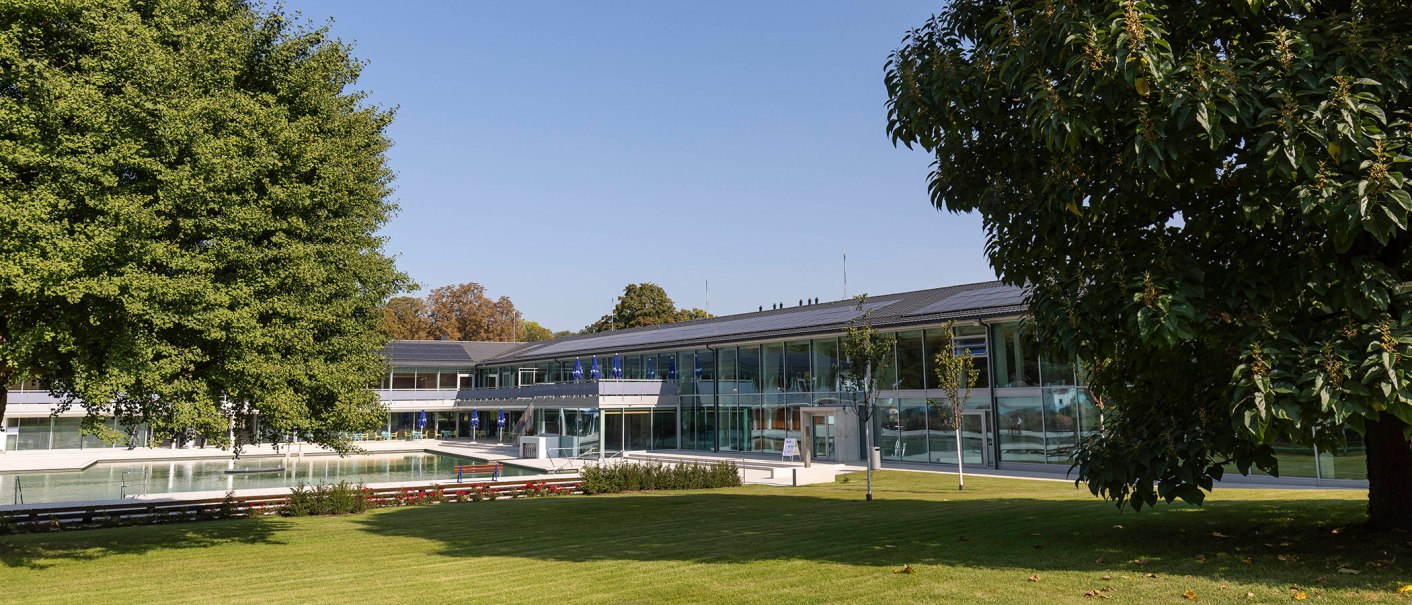 The picture shows the outdoor area of the Mineralbad Berg with a pool, surrounded by modern buildings and green trees under a clear sky., © Stuttgarter Bäder