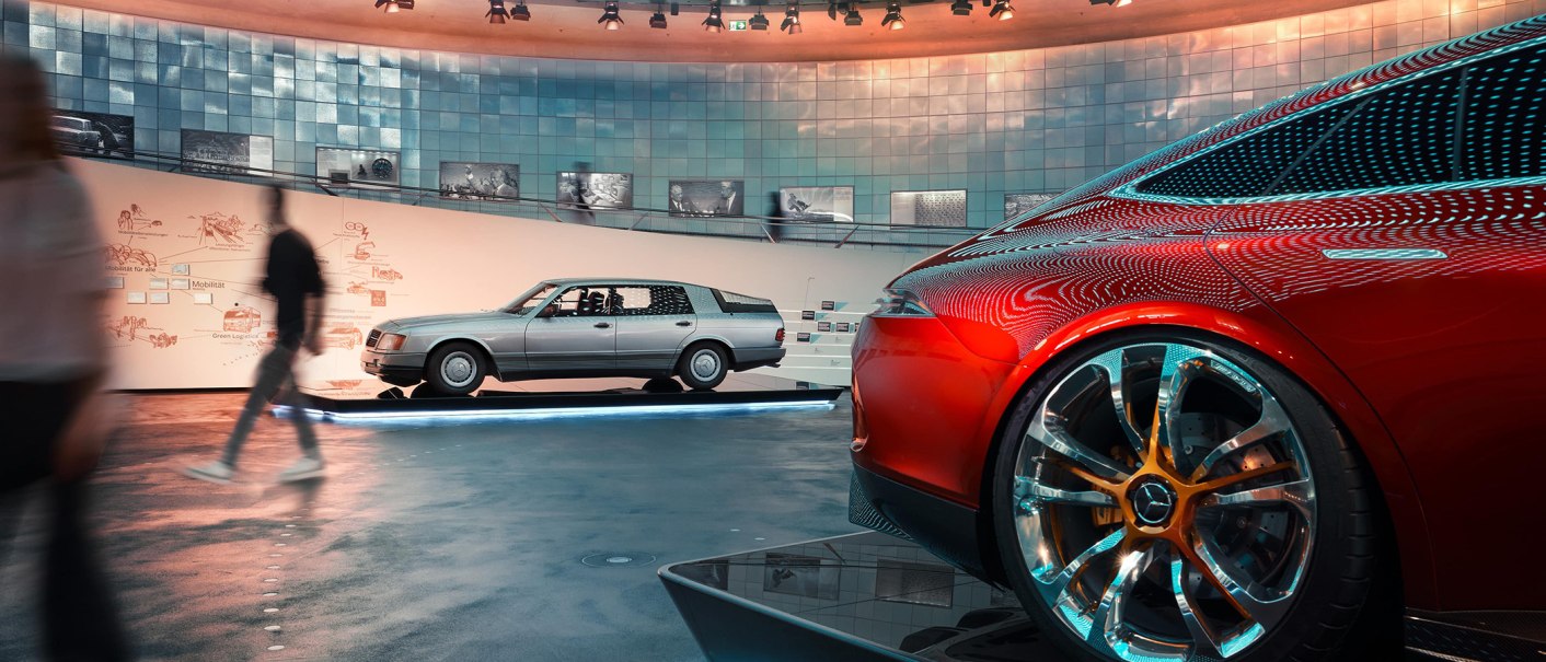 Innenansicht des Mercedes-Benz Museums mit einem roten und einem silbernen Auto. Besucher gehen vorbei, moderne Architektur und Beleuchtung im Hintergrund., &copy; Mercedes-Benz Heritage GmbH
