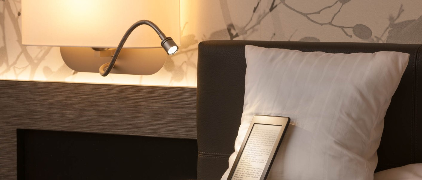 A hotel room with a reading lamp on the wall, a cushion and an e-reader on the bed. The wallpaper has a subtle pattern., &copy; MASEVEN Stuttgart SI-Centrum