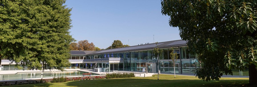The picture shows the outdoor area of the Mineralbad Berg with a pool, surrounded by modern buildings and green trees under a clear sky., © Stuttgarter Bäder