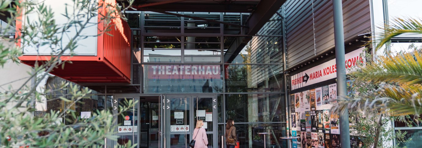 Eingang des Theaterhauses Stuttgart mit rotem Boden, Glasfassade und Plakaten. Zwei Personen betreten das Geb&auml;ude. Pflanzen und moderne Architektur sind sichtbar., &copy; Stuttgart-Marketing GmbH, wpsteinheisser