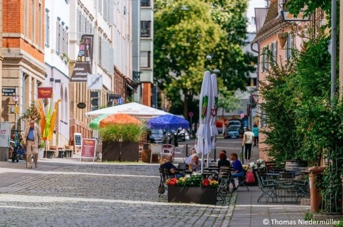 Belebte Straße mit Cafés, bunten Sonnenschirmen und Menschen, die draußen sitzen. Pflastersteine und grüne Pflanzen säumen den Weg., © Stuttgart Marketing GmbH Belebte Straße mit Cafés, bunten Sonnenschirmen und Menschen, die draußen sitzen. Pflastersteine und grüne Pflanzen säumen den Weg., © Stuttgart Marketing GmbH