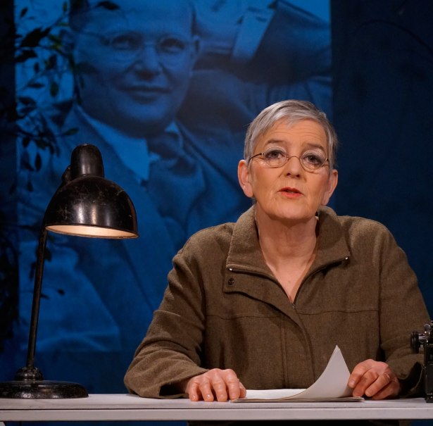 Eine Frau sitzt an einem Tisch mit einer Lampe und hält ein Papier. Im Hintergrund ist ein großes Porträt eines Mannes zu sehen., © © Katja Ritter Eine Frau sitzt an einem Tisch mit einer Lampe und hält ein Papier. Im Hintergrund ist ein großes Porträt eines Mannes zu sehen., © © Katja Ritter