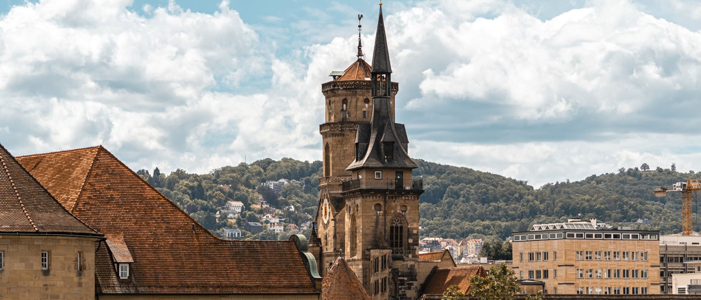 Die Stiftskirche mit ihrem markanten Turm erhebt sich über die Dächer der Stadt, umgeben von grünen Hügeln und einem bewölkten Himmel., © Stuttgart-Marketing GmbH, Sarah Schmid Die Stiftskirche mit ihrem markanten Turm erhebt sich über die Dächer der Stadt, umgeben von grünen Hügeln und einem bewölkten Himmel., © Stuttgart-Marketing GmbH, Sarah Schmid