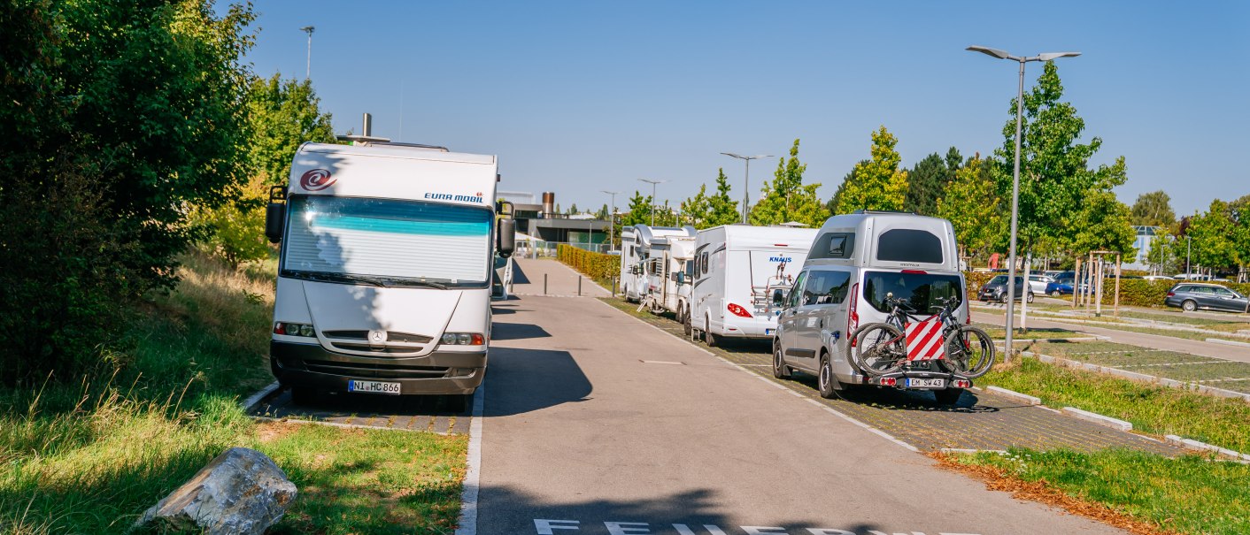 Wohnmobilstellplatz in Fellbach mit geparkten Wohnmobilen und Autos, umgeben von B&auml;umen und klarem Himmel. Auf der Stra&szlig;e steht 'Feuerwehrzufahrt'., &copy; Stuttgart-Marketing GmbH, Thomas Niederm&uuml;ller