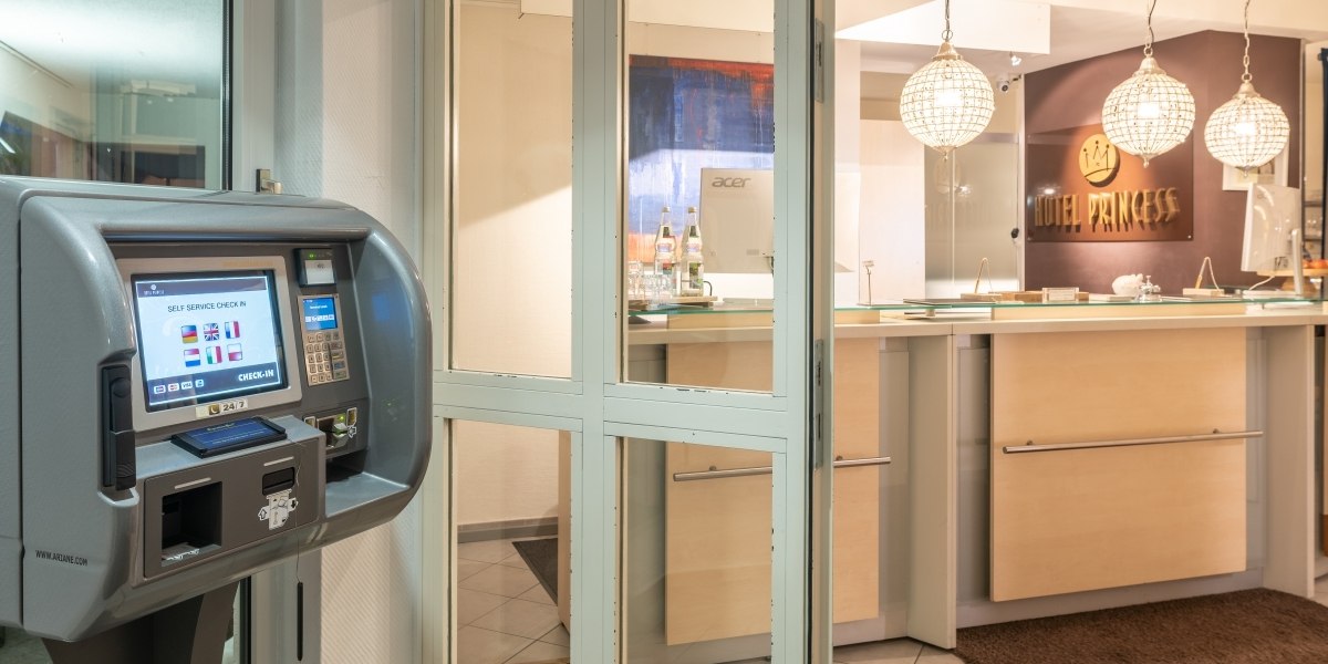 Hotelrezeption mit Selbstbedienungs-Check-in-Terminal und beleuchtetem Empfangstresen. Im Hintergrund das Schild 'Hotel Princess'., © Hotel Princess Hotelrezeption mit Selbstbedienungs-Check-in-Terminal und beleuchtetem Empfangstresen. Im Hintergrund das Schild 'Hotel Princess'., © Hotel Princess