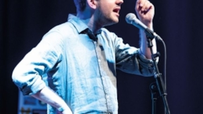 A man in a blue shirt gesticulates into a microphone on a stage., © Altes Schauspielhaus und Komödie im Marquardt e.V. A man in a blue shirt gesticulates into a microphone on a stage., © Altes Schauspielhaus und Komödie im Marquardt e.V.